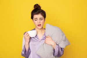 sleepy brunette female in silk pajamas with cup of coffee and pillow yawning
